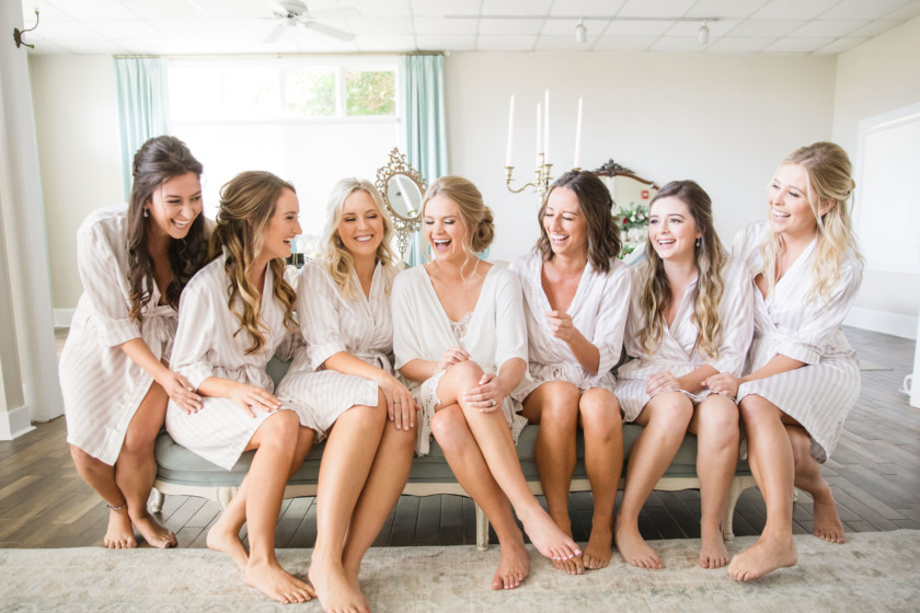 bride and bridesmaids laughing in robes sitting on couch in bridal suite at white room