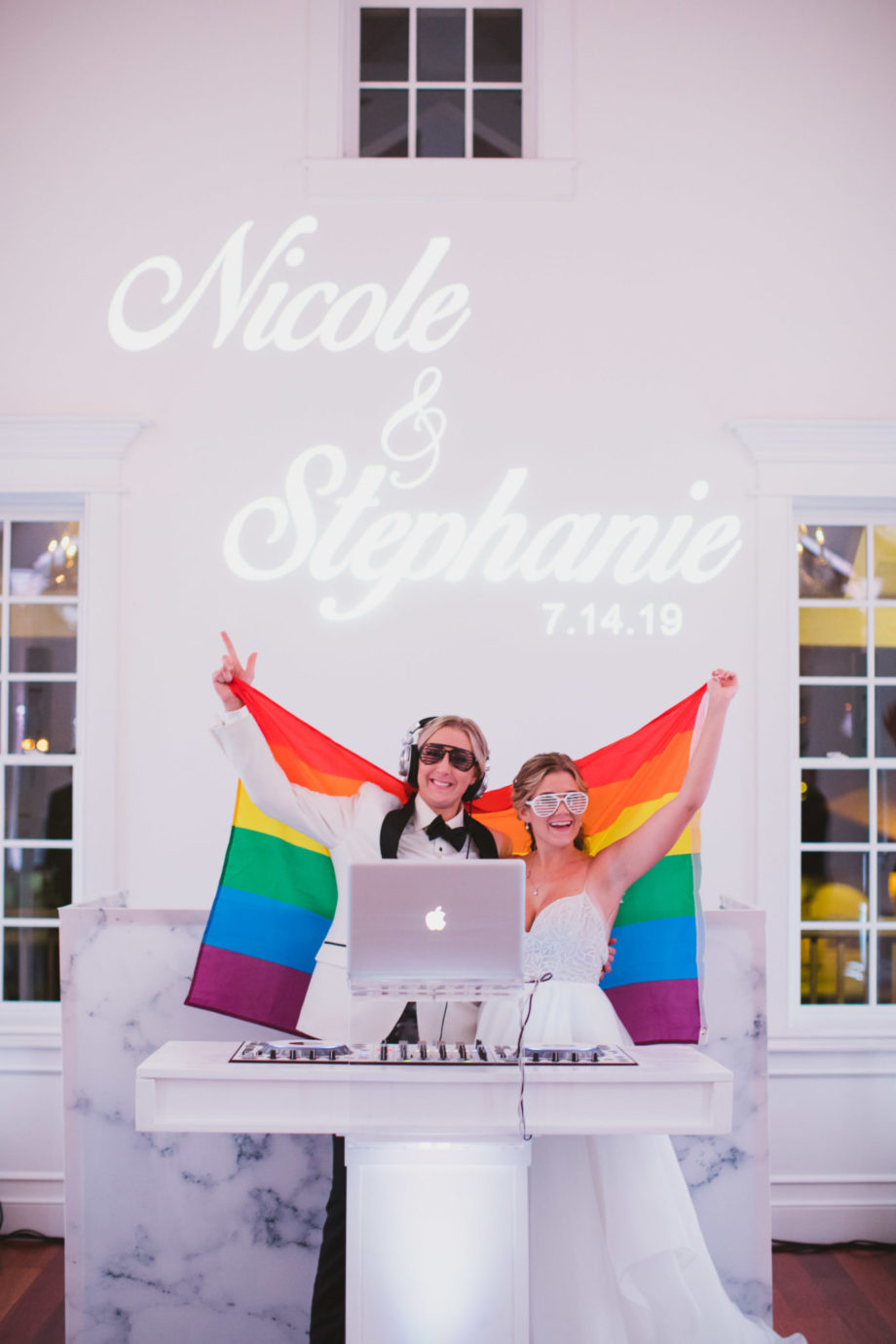 two brides holding up the gay pride flag and djing at their wedding reception at white room