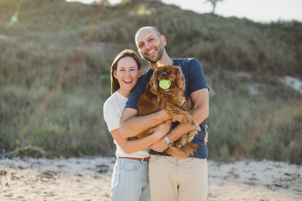 couple with their dog holding a ball in his mouth on the beach