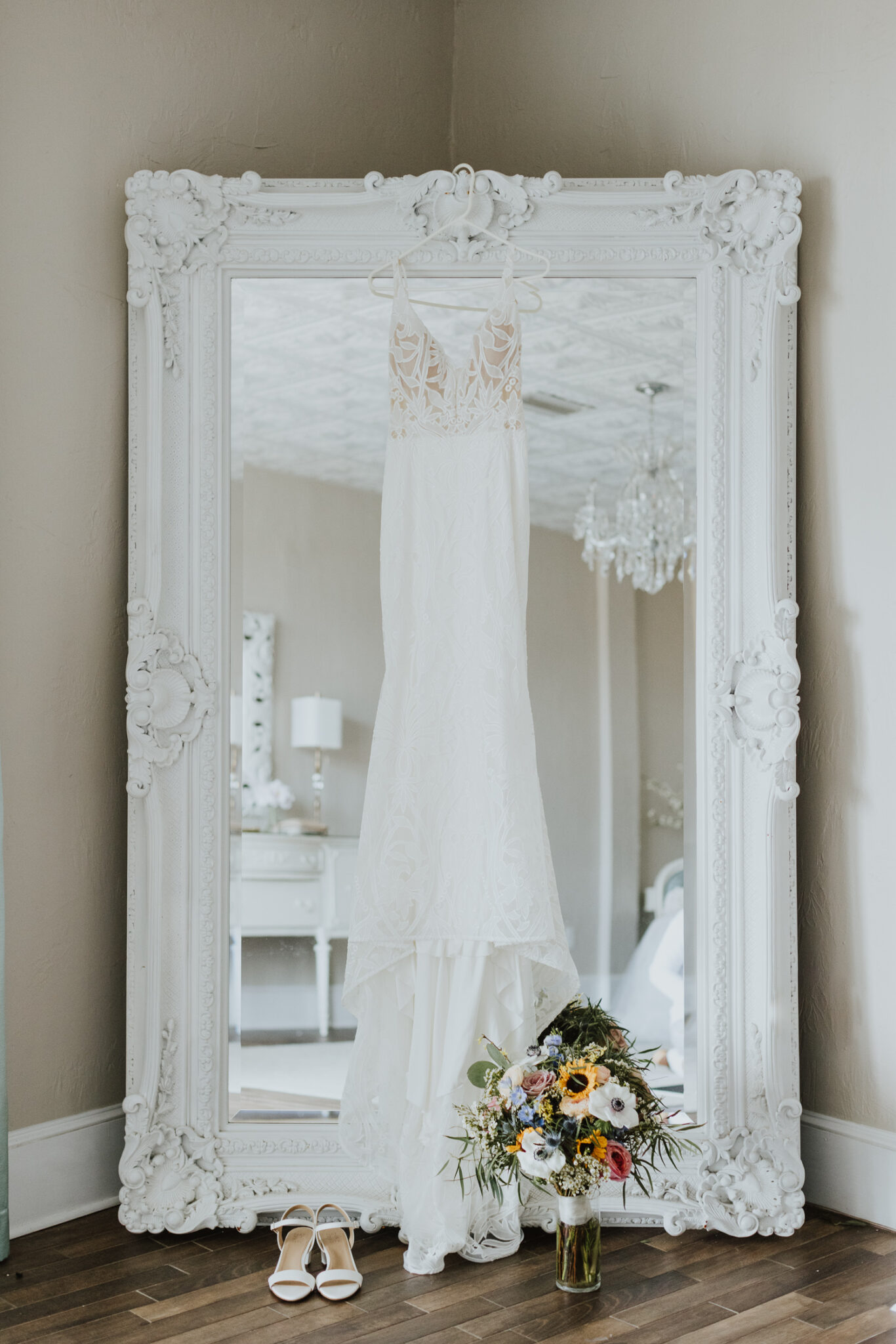 bridal gown hanging on white room bridal suite large mirror with shoes and bridal bouquet on the floor in front of dress