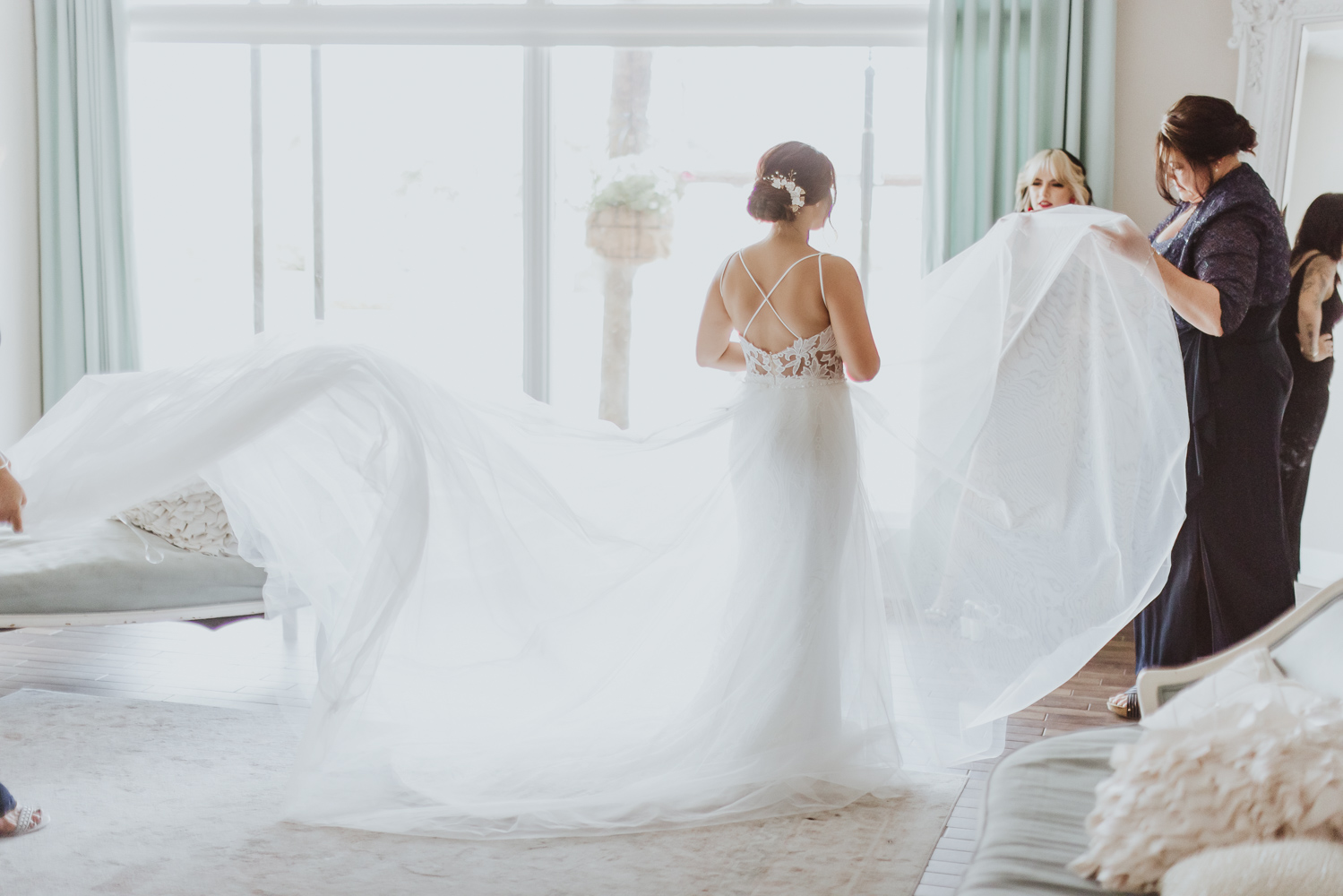 bride standing in white room bridal suite with moms helping fluff out her dress 