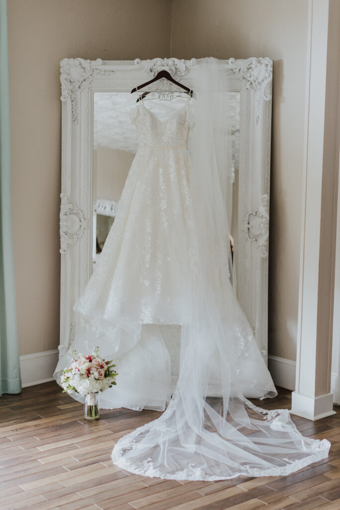 bridal gown and veil hanging on floor length mirror with bouquet on floor of bridal suite of the white room in st. augustine