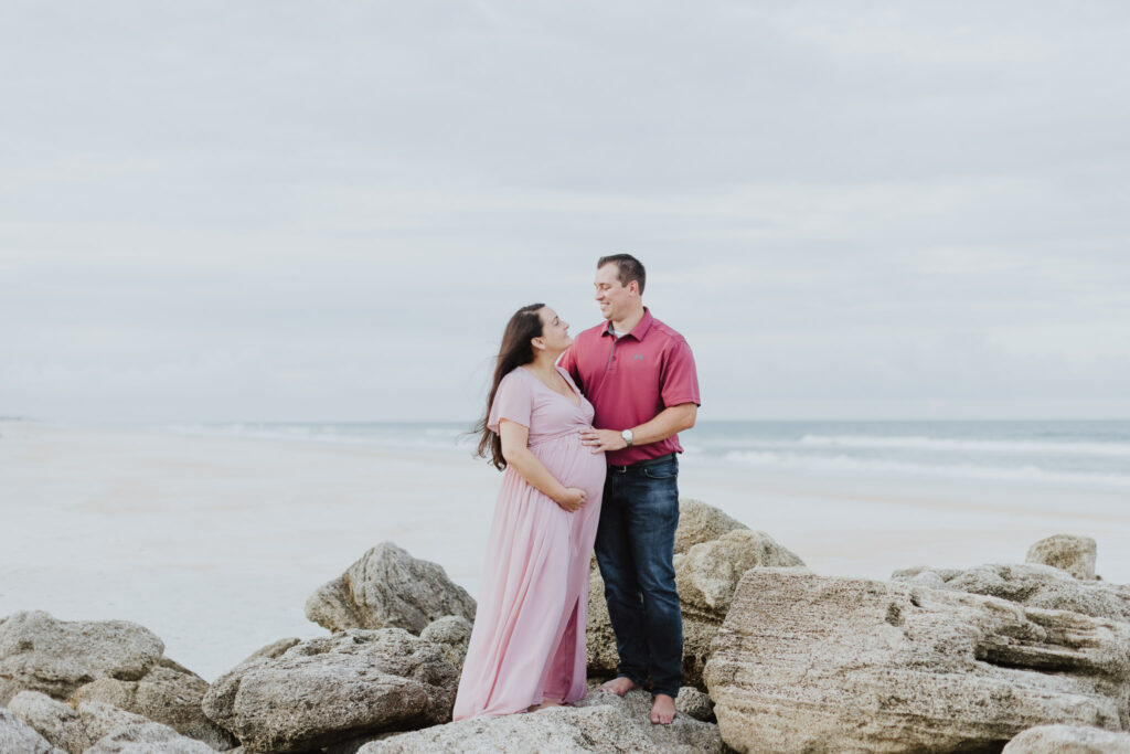 couple looking at each other standing on rocks on beach at washington oaks touching baby belly during maternity photo session