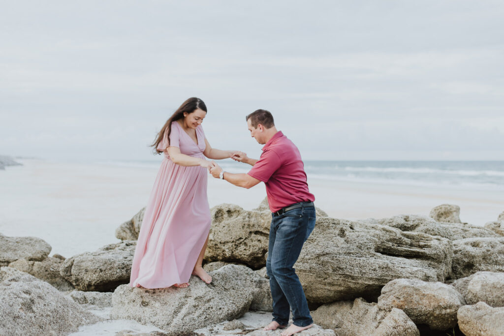 man helping wife hop down from rock along the washington oaks park beach during maternity session