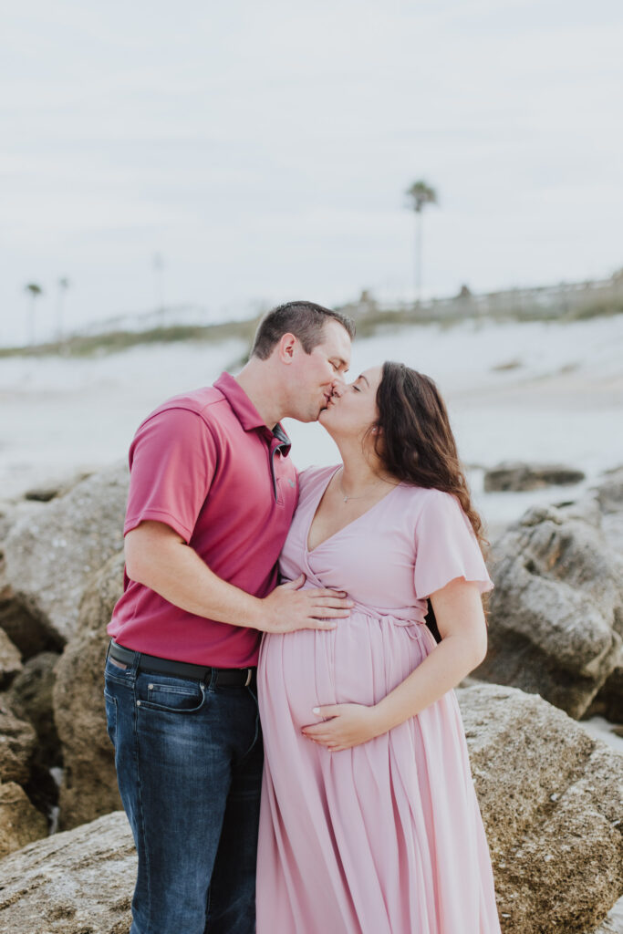 soon to be parents kissing while standing in front of rocks at washington oaks park beach during maternity session 