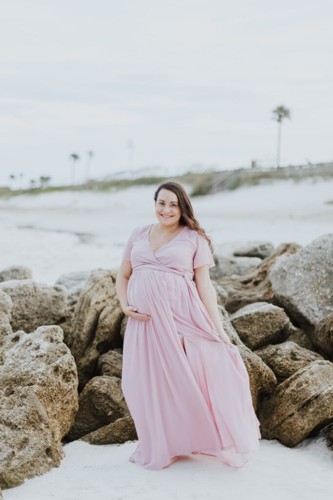 mom to be holding baby belly wearing pink flowing dress standing on beach at washington oaks park beach side