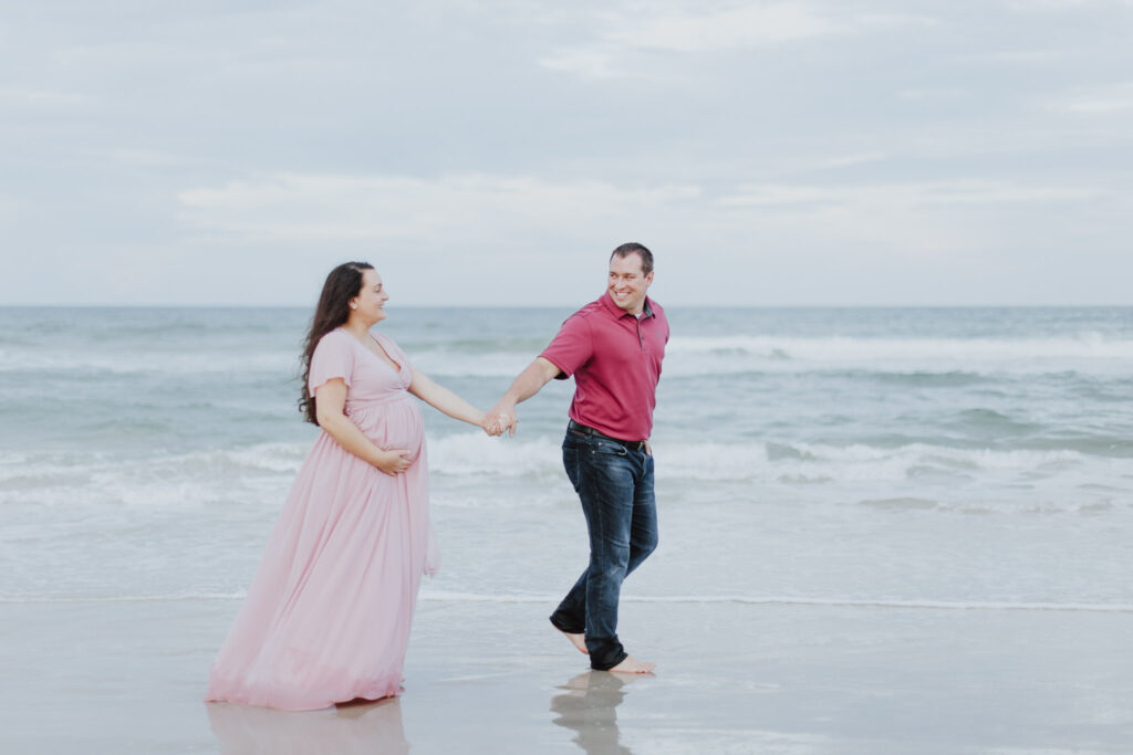 couple holding hands and walking along water at beach during maternity session 