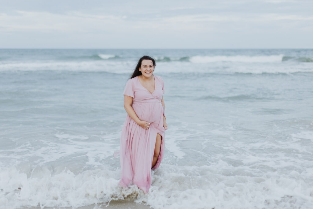 mom to be holding belly while putting feet in ocean waves during maternity session