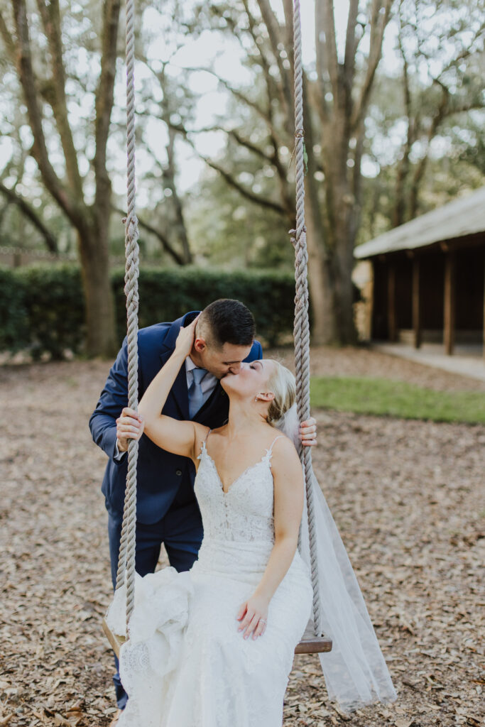 bride sitting on rope swing hanging from large oak tree and groom kissing her