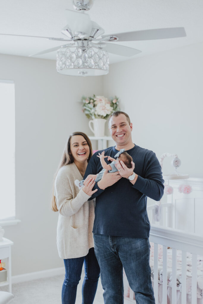 parents laughing while holding newborn baby girl and stand in nursery