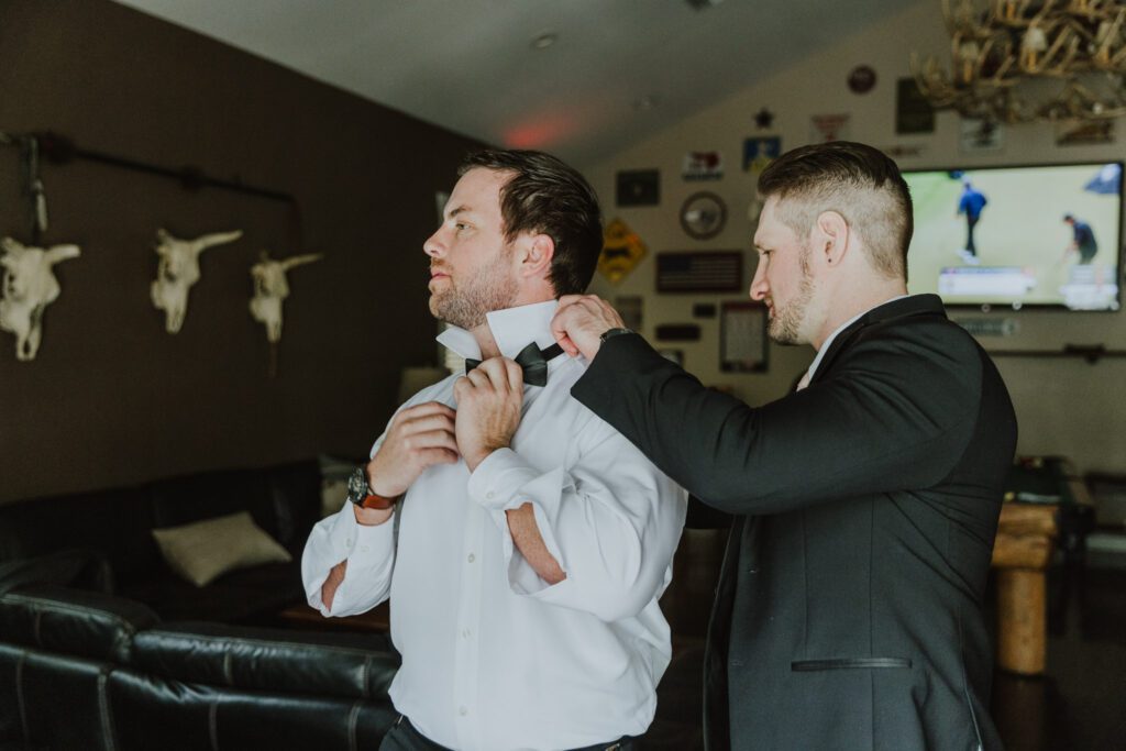 best man helping groom put bow tie on while getting ready in the groom's suite of bowing oaks wedding venue in jacksonville