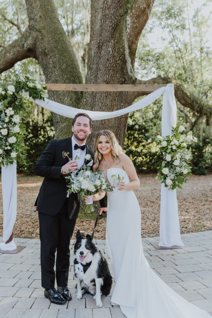 groom and bride holding drinks in hand and posing with their dog on their wedding day in jacksonville, fl