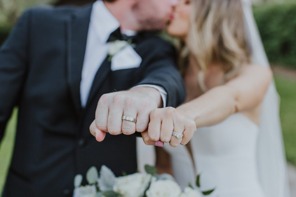 the bride and groom holding their hands as fists next to one another show their wedding bands and kissing in the background