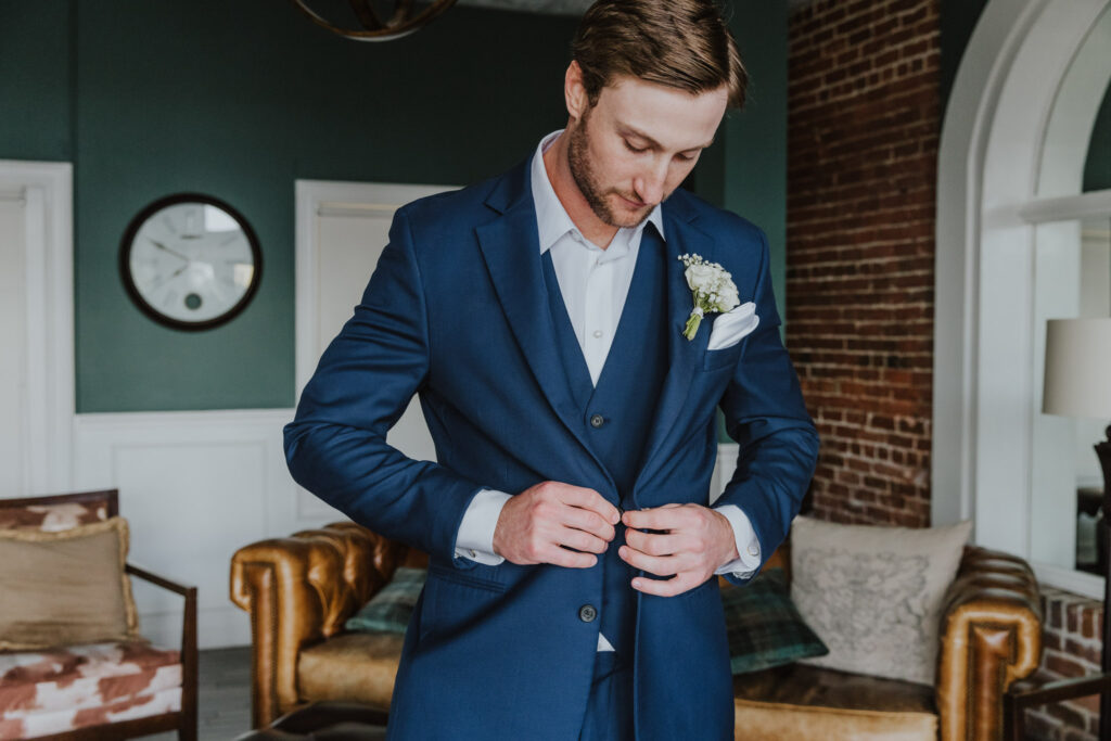 groom bottoning up navy suit while standing in white room groom's suite in st. augustine