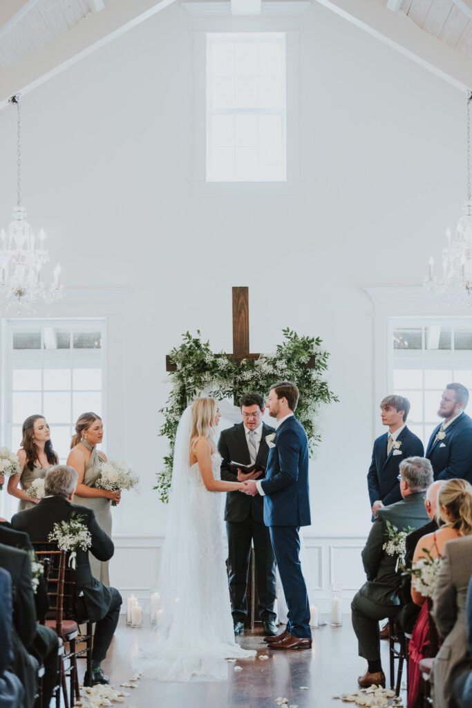bride and groom holding hands looking at each other during their villa blanca ceremony with wood cross in back of them decorated with greenery and white florals