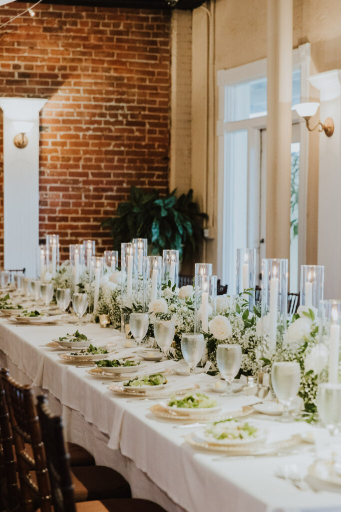 long wedding reception table setup with tall white candles in glass, white florals and greenery