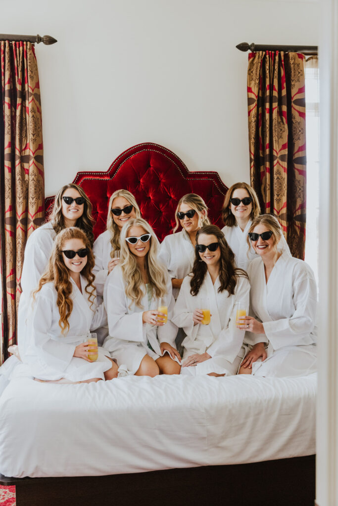 bride with bridesmaids sitting on casa monica bed in white robes, holding mimosas in hands and wearing sunglasses