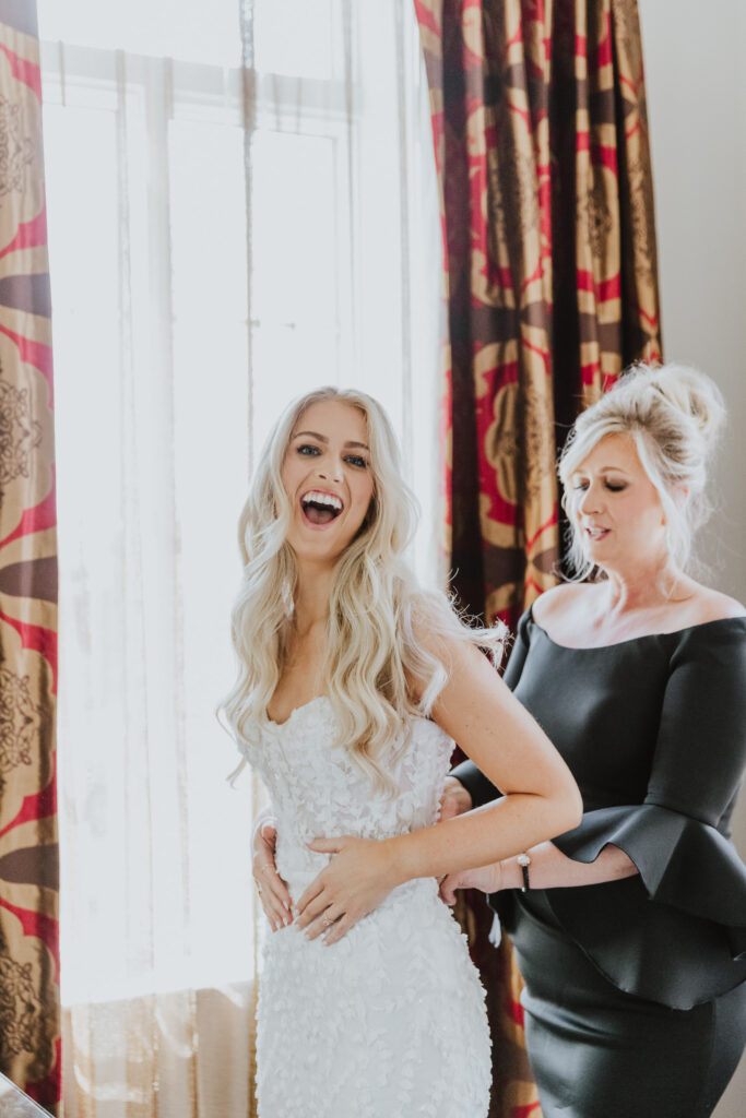 bride laughing while mom is zipping up her bridal gown standing in casa monica hotel room getting ready
