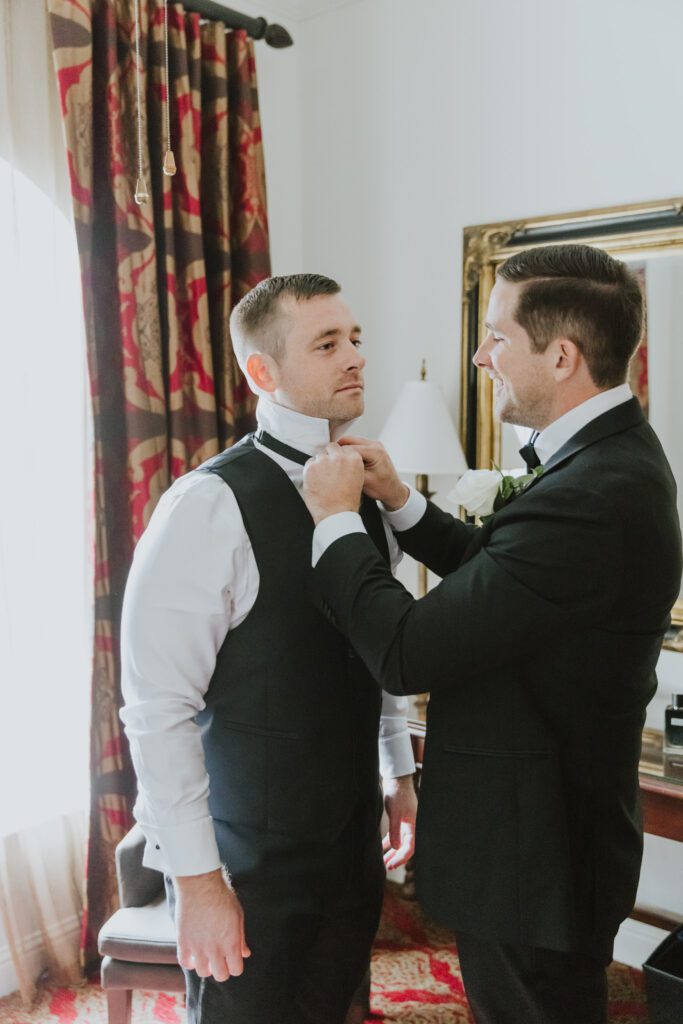 groom getting ready with best man adjusting his bowtie before wedding at the white room