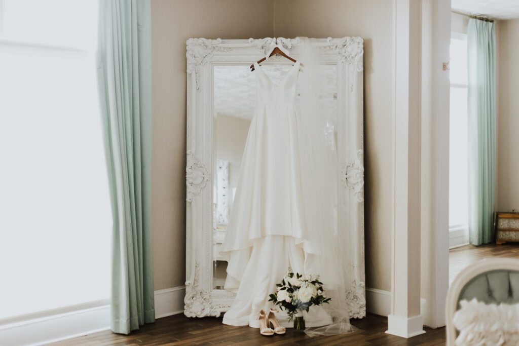 bride's wedding gown hanging on large white mirror with bouquet and shoes in front hanging in white room bridal suite
