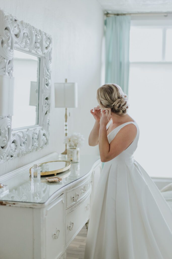 bride standing at mirror in the bridal suite of the white room putting on her earring