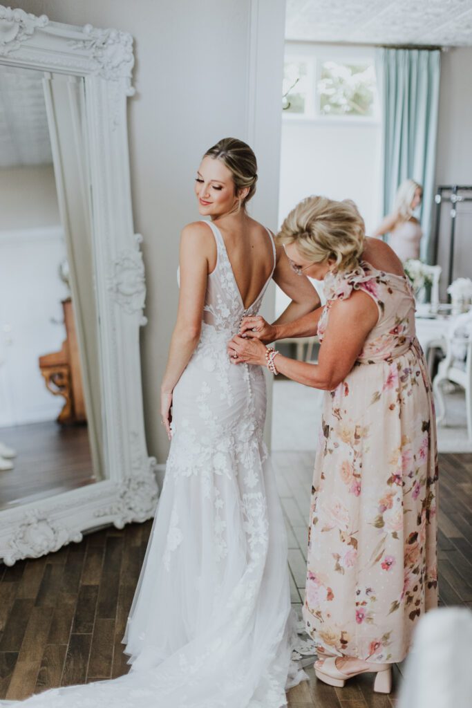 bride's mom buttoning up the back of bride's gown while she stands in front of mirror in the white room bridal suite