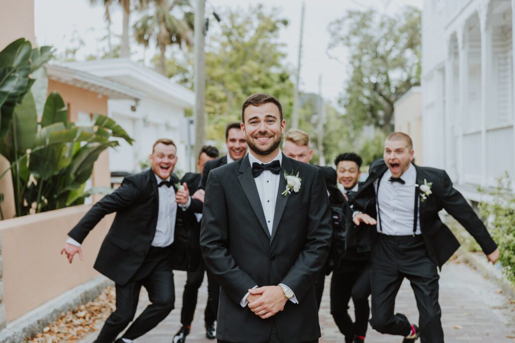groom standing in street with groomsmen running up behind him 