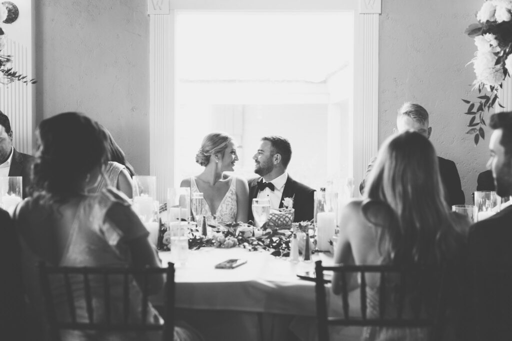 bride and groom sitting at the head table at their reception at the white room looking at each other