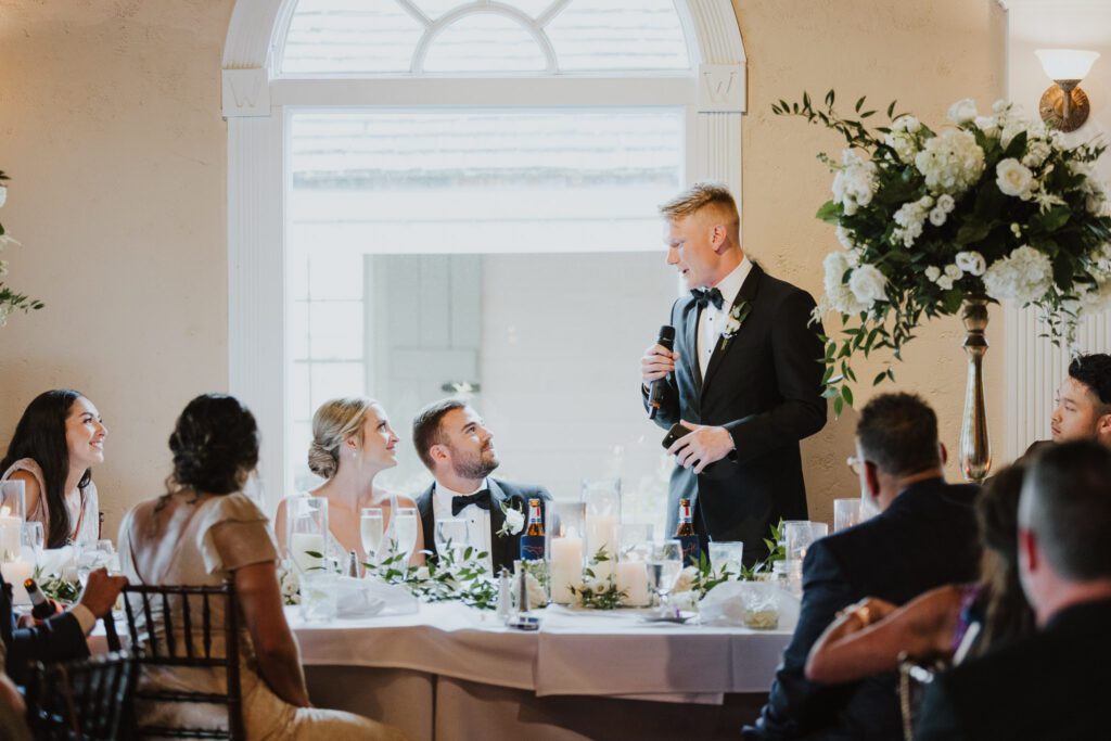 best man looking at the bride and groom and giving speech during white room wedding reception 