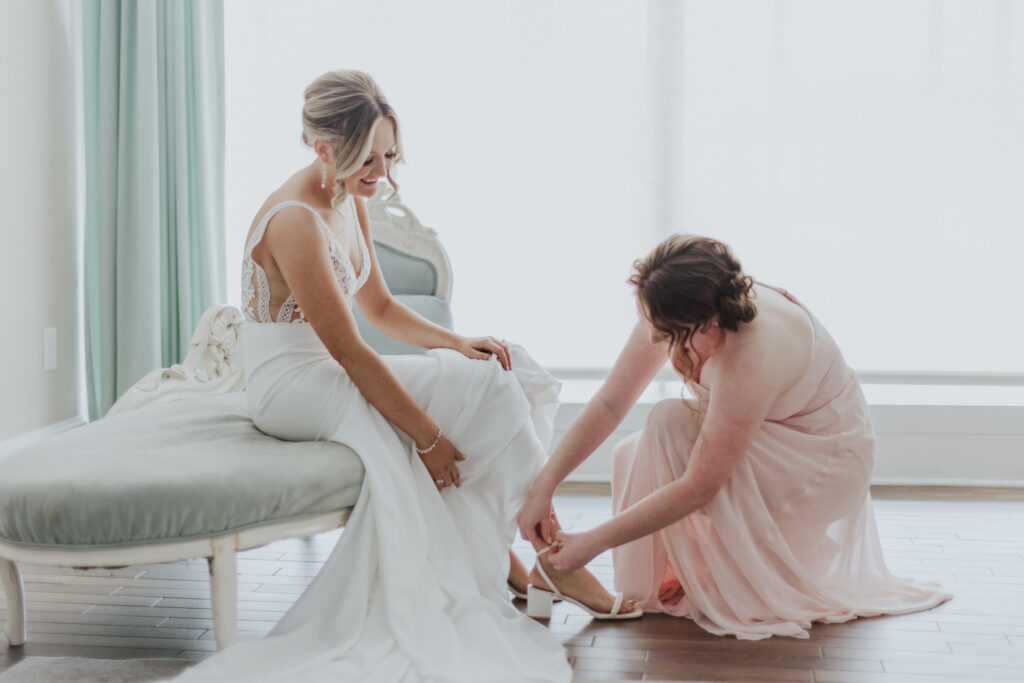 bride sitting down and bridesmaid helping her put her shoes on while getting dressed at the white room bridal suite.